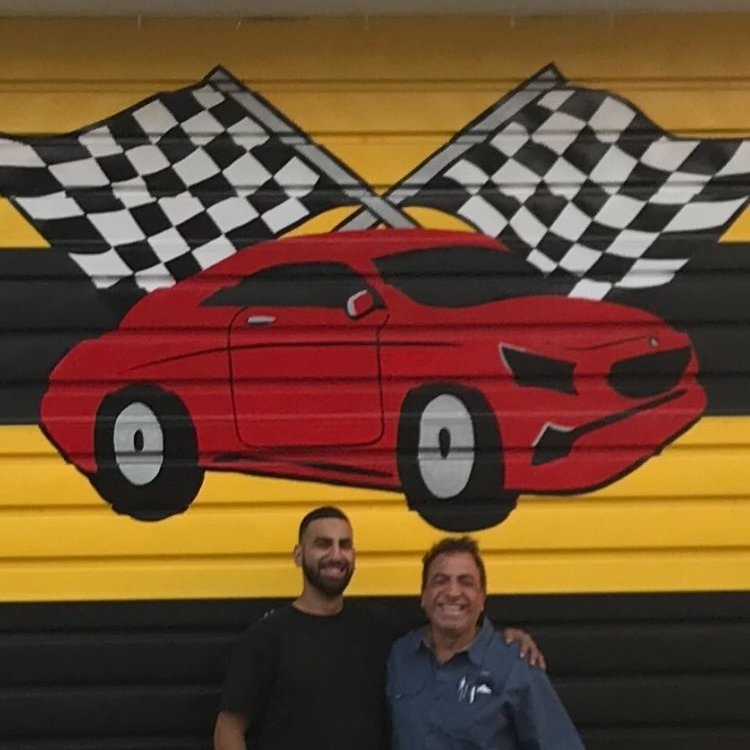 Father and son owners of Perfect Lube Car Care standing in front of the checkered flag mural